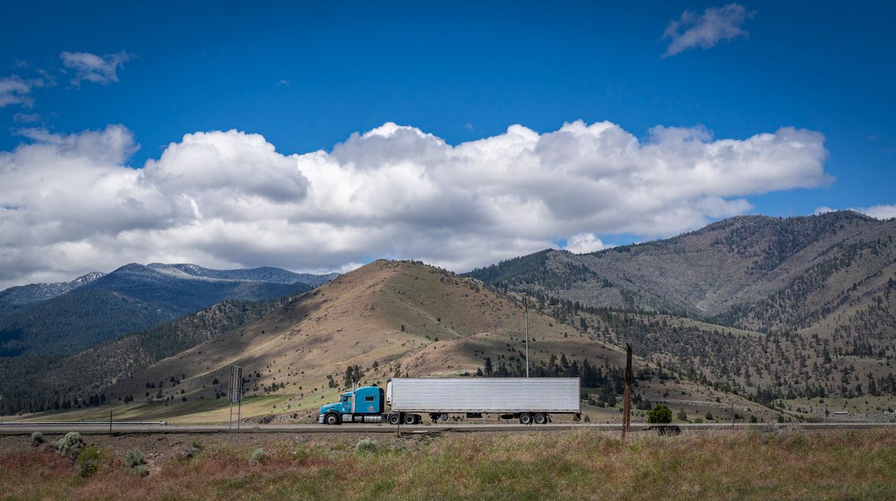A blue semi-truck traveling beneath cloudy skies on a mountain highway